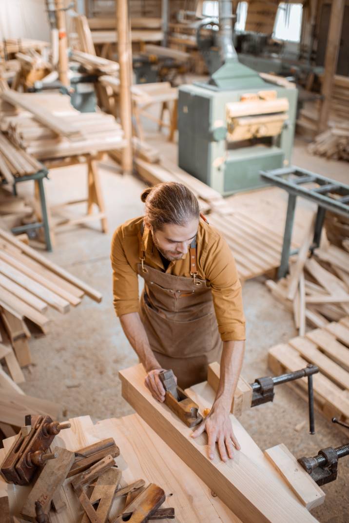 Handsome carpenter working with a wood, planing a bar with a plane on the worbench in the carpentry workshop. View from above
